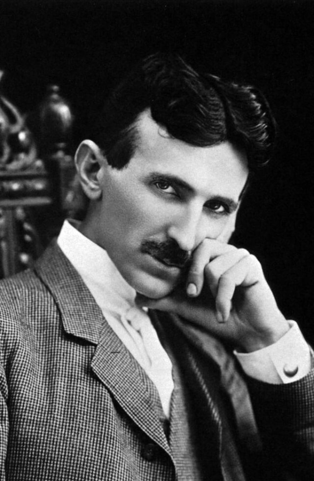 A black-and-white portrait of a man with dark hair and a mustache, wearing a suit. He is seated, resting his head on his hand, and gazing thoughtfully at the camera against a dark background.