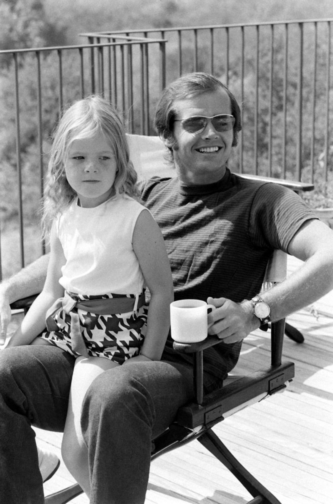 A man wearing sunglasses smiles while sitting in a chair outdoors, holding a mug. A young girl with light hair in a sleeveless top and patterned shorts sits on his lap, looking away. There is a railing and foliage in the background.