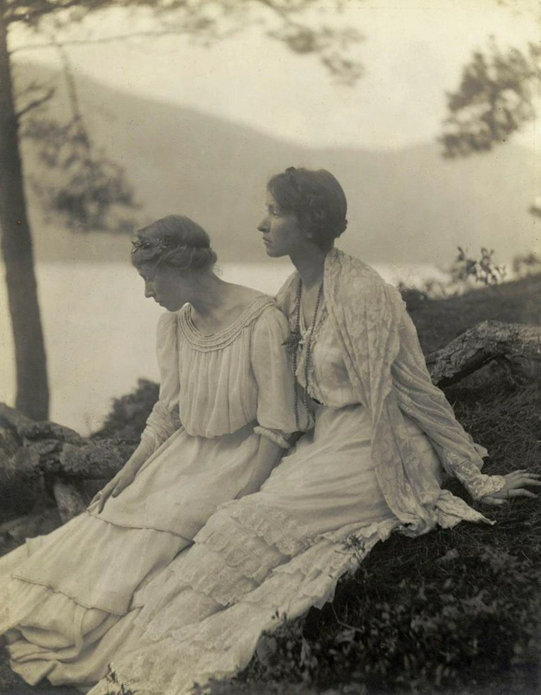 Two women in light, flowing dresses sit close together on a grassy hillside near a lake, with trees and distant hills in the background. The image has a soft, vintage appearance.