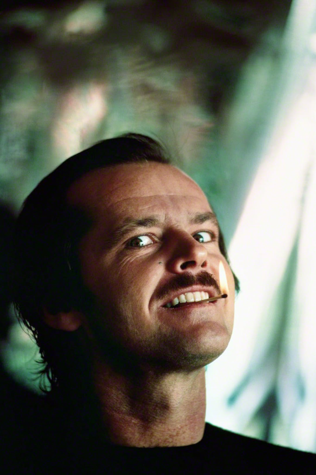A man with slicked-back hair and a mustache grins mischievously, holding a lit matchstick between his teeth. The background is blurred with green and white tones, creating a dramatic and intense atmosphere.
