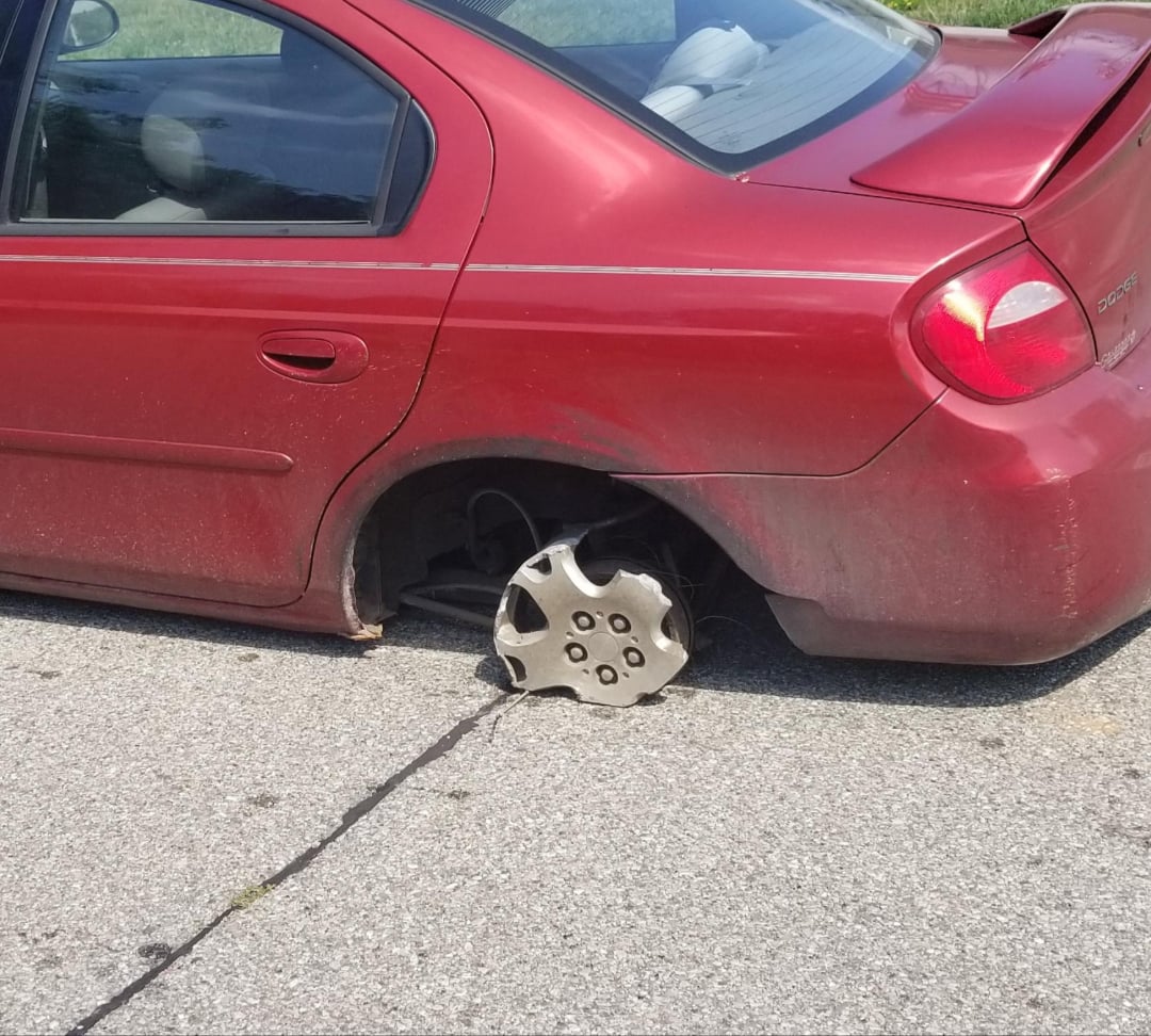 A red car is parked on the street with its rear left tire missing, exposing the metal rim and wheel cover lying on the ground nearby. The car's rear is low to the pavement.