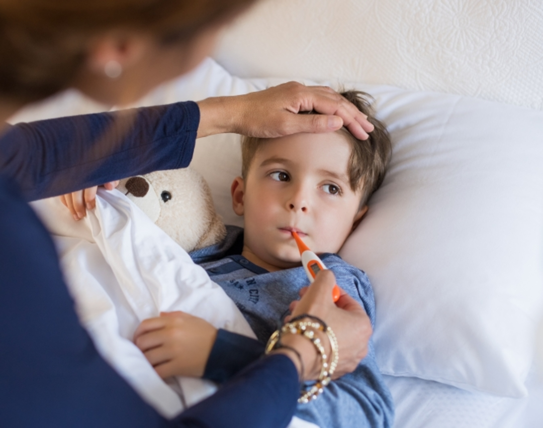 A young boy lies in bed with a thermometer in his mouth, holding a teddy bear, while an adult checks his forehead for a fever.