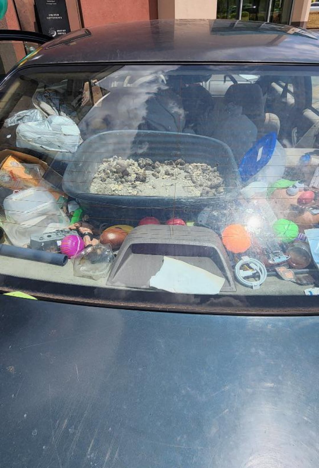 A car seen from the rear window is cluttered with various items, including a cat litter box filled with litter and numerous colorful toys and objects scattered across the backseat and rear deck.