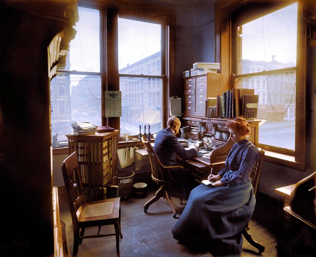 A man and a woman in early 20th-century clothing work at desks in a sunlit office with large windows, wooden furniture, shelves of books, and papers scattered on the desks. Buildings are visible outside the windows.
