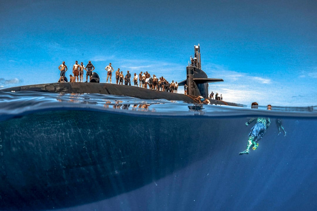 A group of people stand on top of a surfaced submarine in the ocean, while a swimmer in flippers is visible underwater near the vessel. The bright sky contrasts with the deep blue water.