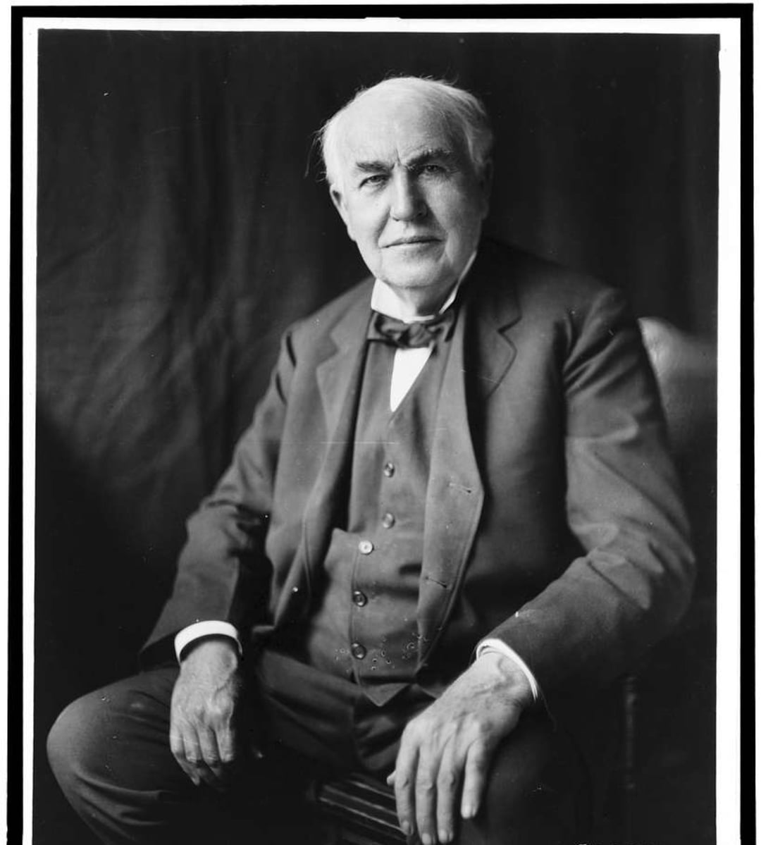 An elderly man with thinning hair sits in a suit and bow tie, facing the camera with a slight smile, against a plain dark background.