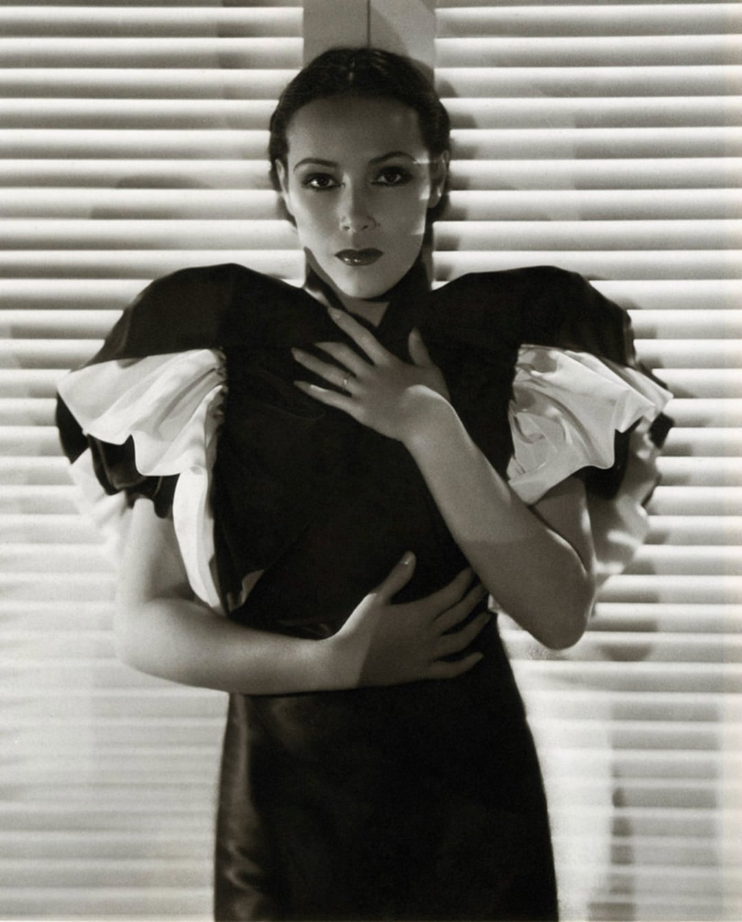 A woman in a dramatic black dress with large, ruffled sleeves stands in front of closed window blinds, one hand on her chest and the other on her waist, looking directly at the camera in a classic black-and-white portrait.