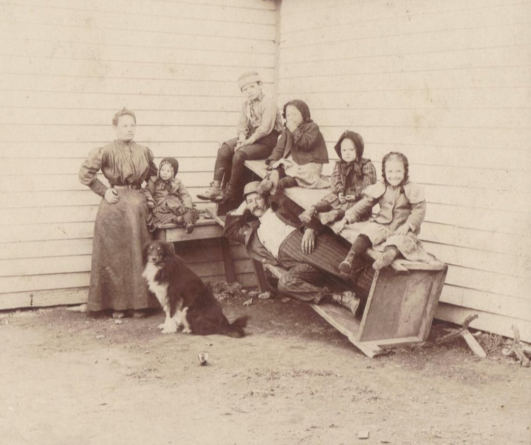 A sepia photo of a family by a wooden wall: a woman stands with hands on hips, five children sit or stand on wooden benches, a man reclines on one bench, and a dog sits in front. All wear old-fashioned clothes.