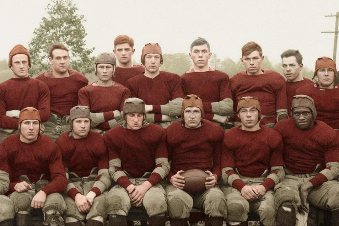 A vintage football team poses for a group photo outdoors, wearing old-fashioned uniforms with padded leather helmets and maroon jerseys. The players sit and stand in two rows, with one holding a football.