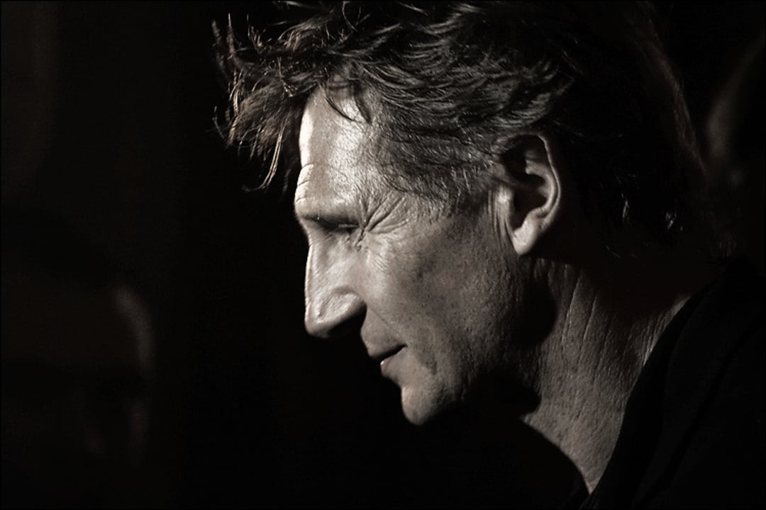 A sepia-toned side profile of a middle-aged man with tousled hair, lit dramatically from the left, showing strong facial features and a contemplative expression against a dark background.