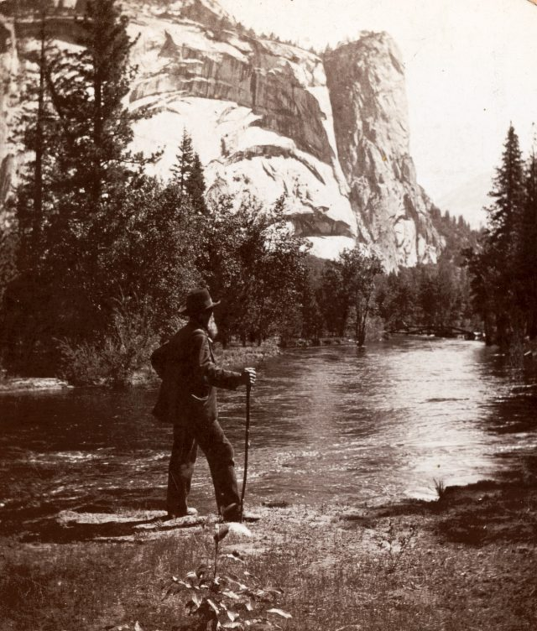 A man with a hat and walking stick stands by a riverbank, looking toward tall pine trees and a large, rocky mountain in the background. The scene is natural and serene, with flowing water and rugged cliffs.