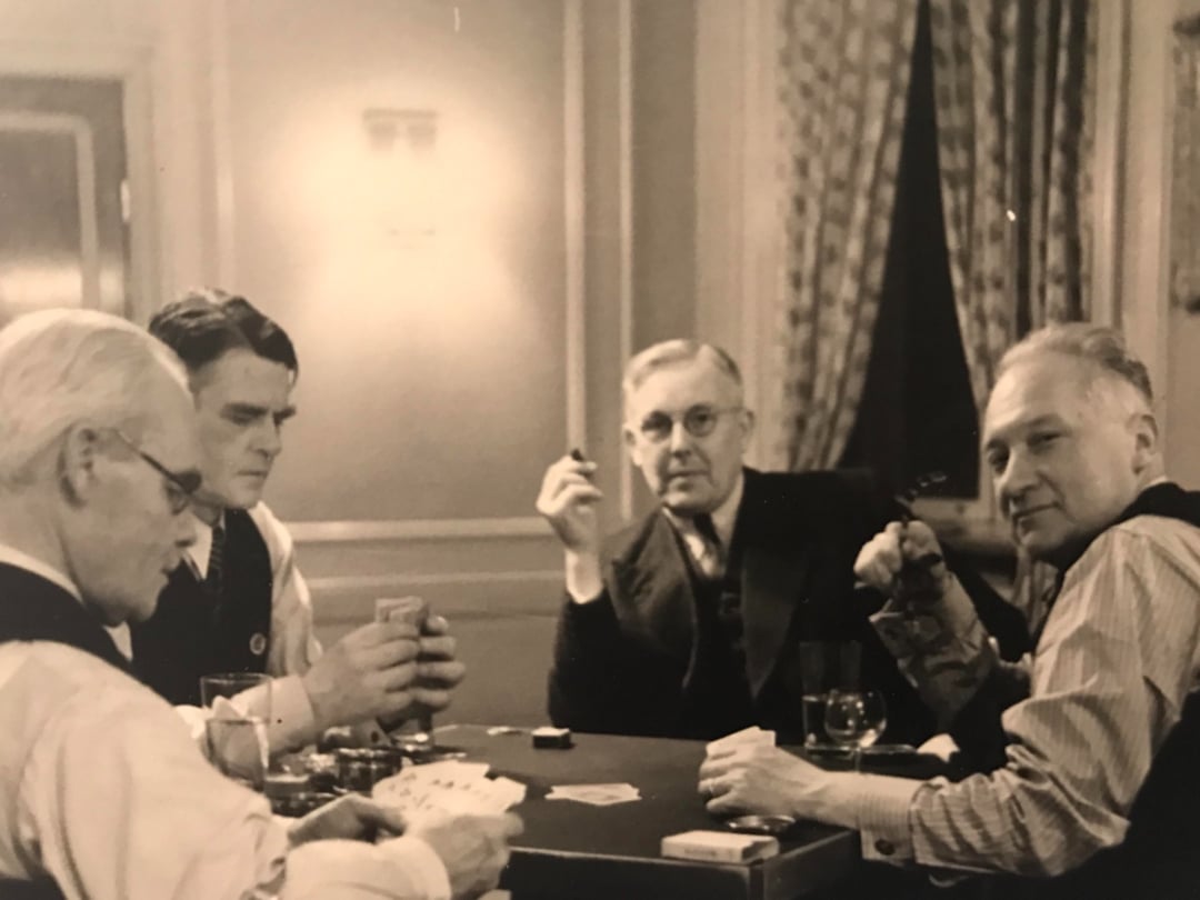 Four men sit around a table playing cards. Some of them hold drinks and cigars, and all appear engaged in the game. The setting is indoors, with curtains and framed windows in the background.