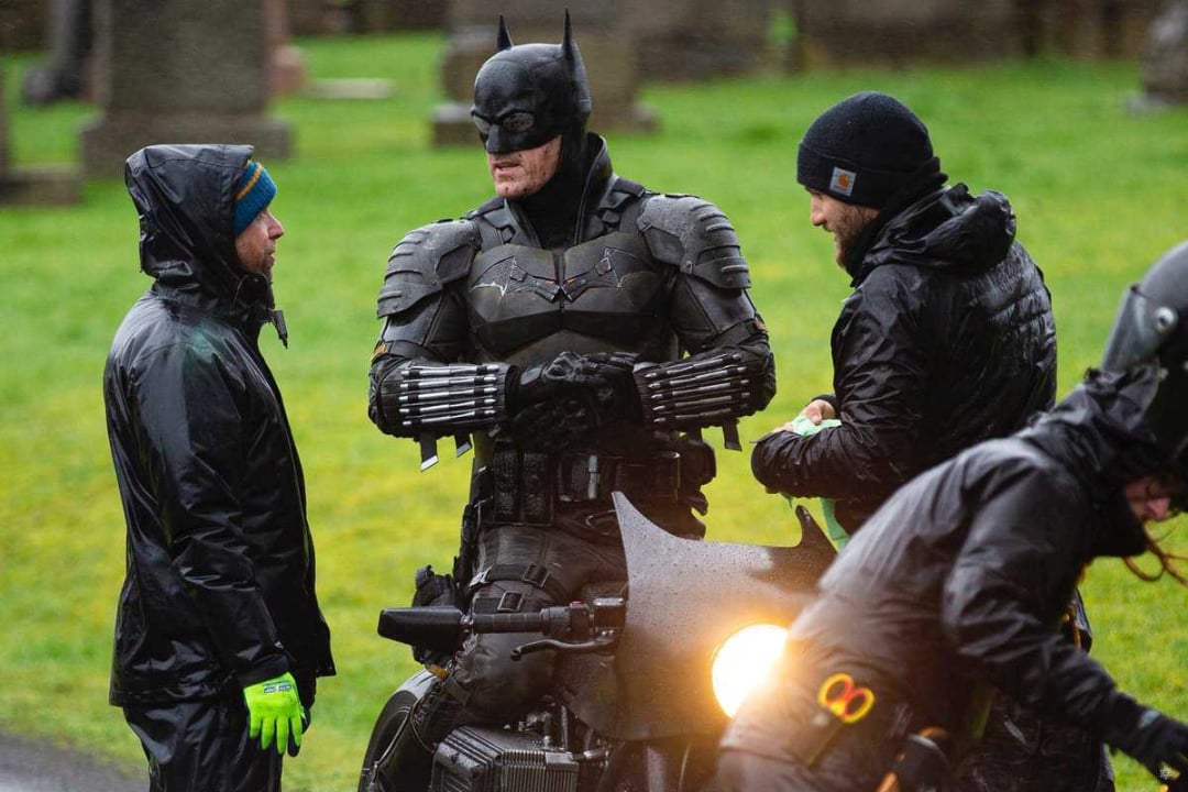 A person dressed as Batman stands beside a motorcycle, talking with two crew members wearing rain jackets and hats on a grassy set, possibly during a film shoot. Another crew member is adjusting something on the motorcycle.