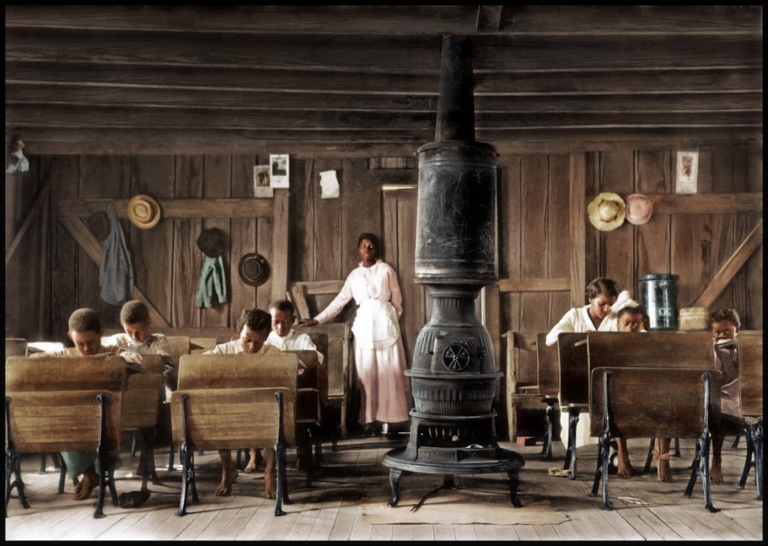 A teacher stands at the front of a rustic wooden classroom with a large stove in the center. Several children sit at wooden desks, focused on their work. Hats and coats hang on the wall behind them.