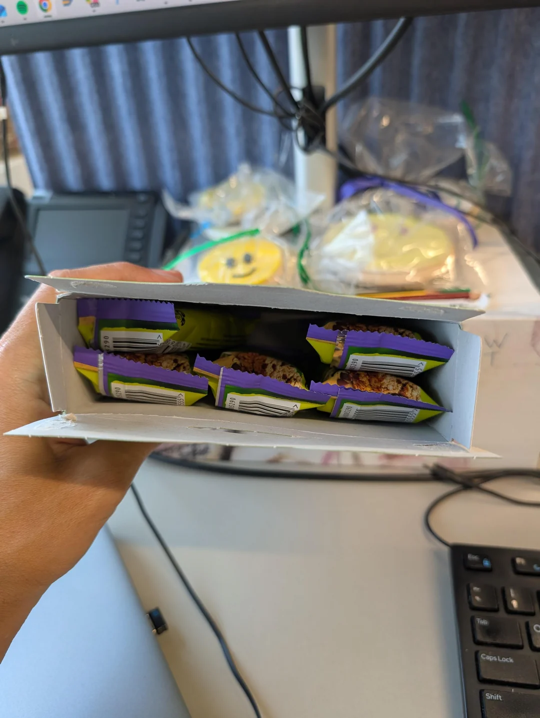 A hand holding an open box containing six individually wrapped granola bars. In the background, decorated cookies and office items are visible on a desk with a computer monitor and keyboard.