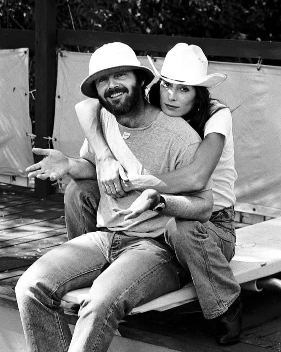A bearded man in a bucket hat and a woman in a cowboy hat sit together outdoors. She hugs him from behind while he sits on a bench, shrugging with a playful expression. Both appear relaxed and casual.