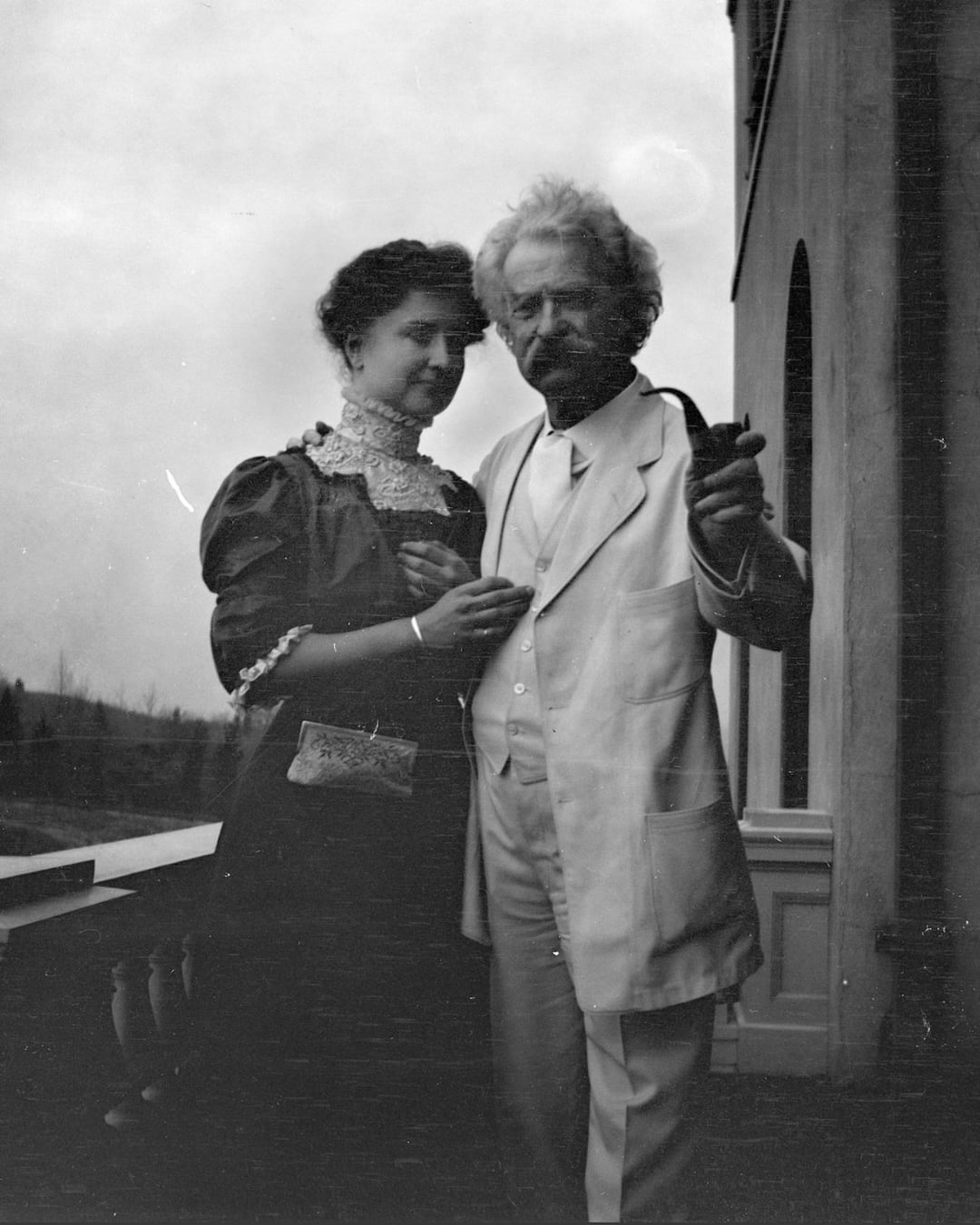 A black-and-white photo of a woman in a dark dress with lace details standing beside an older man in a light suit, who is holding a pipe and pointing, on a terrace with a building and landscape in the background.