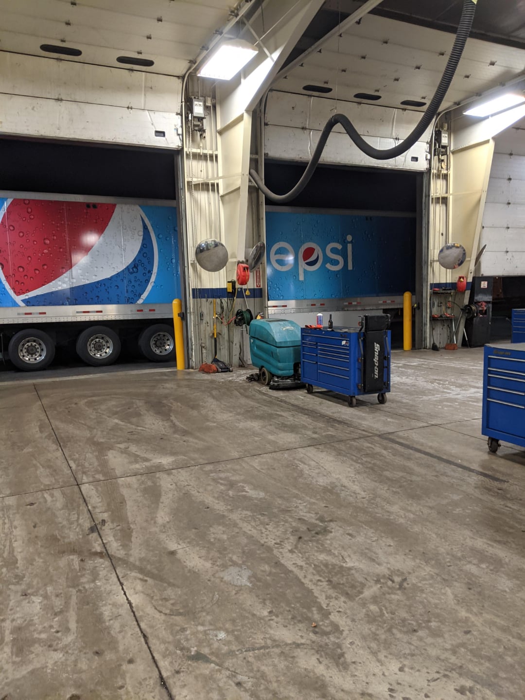 A Pepsi delivery truck is parked inside a large garage with concrete floors. Blue tool chests, cleaning equipment, and industrial fans are visible in the workspace. Overhead lights illuminate the scene.
