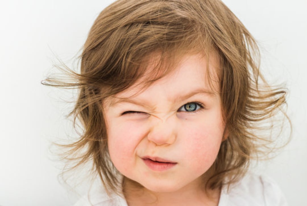 A young child with messy brown hair squints one eye and scrunches their nose, making a playful, mischievous expression against a plain white background.