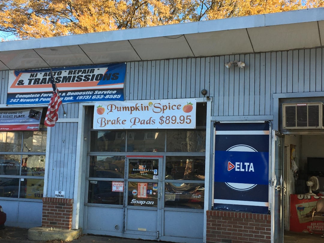 The front of an auto repair shop with signs advertising "Pumpkin Spice Brake Pads $89.95," "NJ Auto Repair & Transmissions," and a blue DELTA sign near the entrance. An American flag is on display.