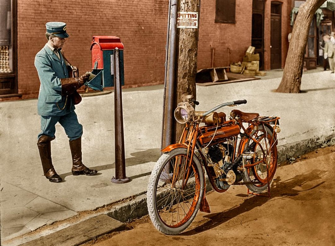 A vintage photo of a mail carrier in a blue uniform collecting letters from a red mailbox on a city sidewalk, with an old-fashioned orange motorcycle parked nearby. A "No Spitting" sign is attached to a pole.