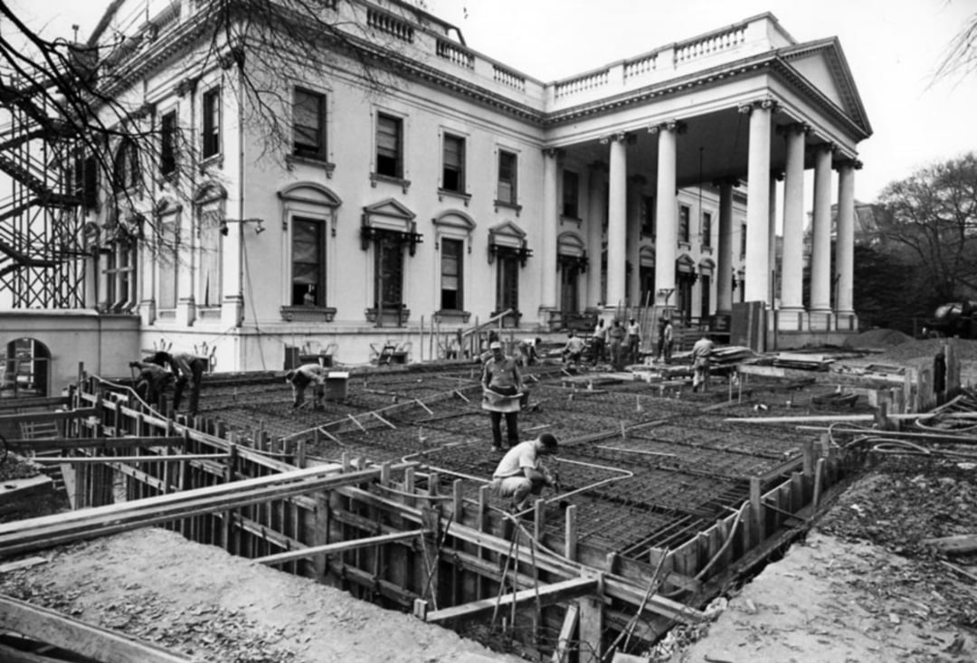 Black and white photo of workers constructing a steel framework for a new floor or foundation in front of a large, classical-style building with tall columns and multiple windows. Trees and scaffolding are visible.