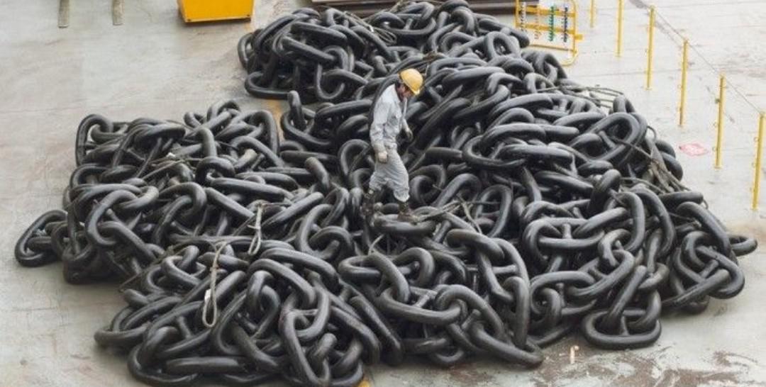 A person in a yellow hard hat and gray work clothes stands on top of a massive pile of thick, heavy black metal chains in an industrial setting with barriers visible in the background.
