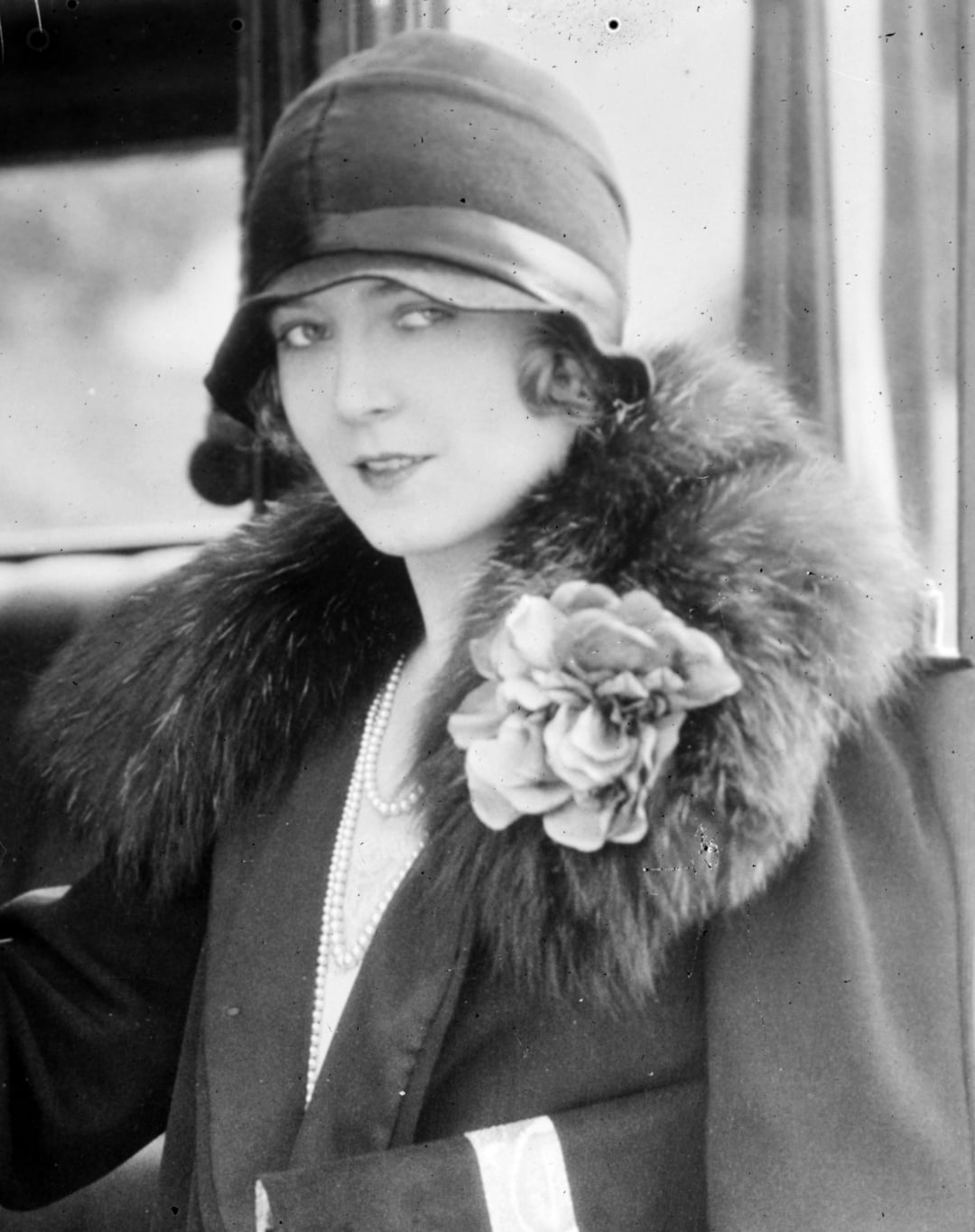 A woman wearing a cloche hat, a fur-collared coat, and a large flower brooch sits beside a car. She has a vintage 1920s style, with wavy hair and layered pearl necklaces.