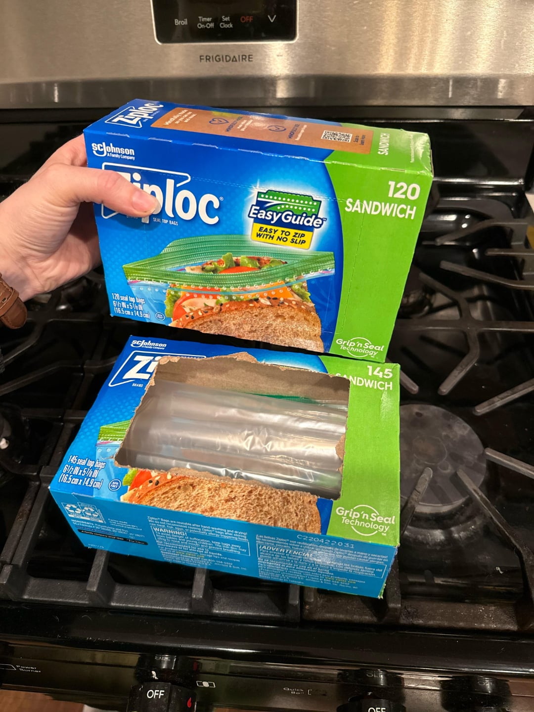 A hand holds a Ziploc sandwich bag box above another open, partially empty Ziploc sandwich bag box on a stove with a stainless steel oven behind them.