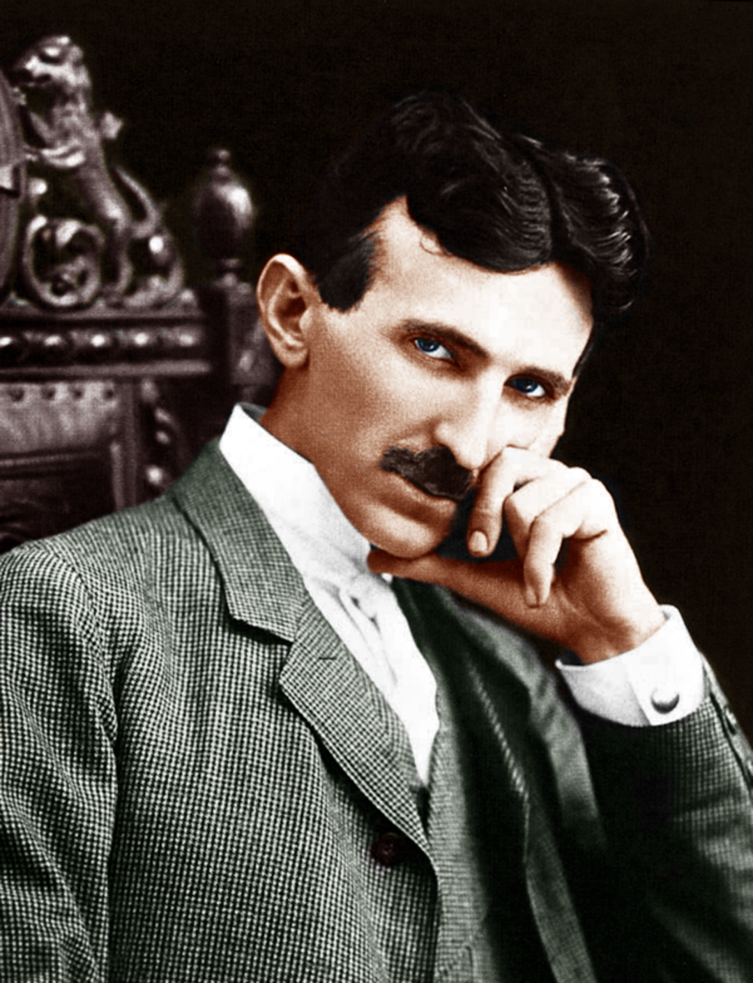 A man with dark hair and a mustache sits on an ornate chair, wearing a green checkered suit and a white shirt, resting his head on his hand and gazing thoughtfully at the camera.