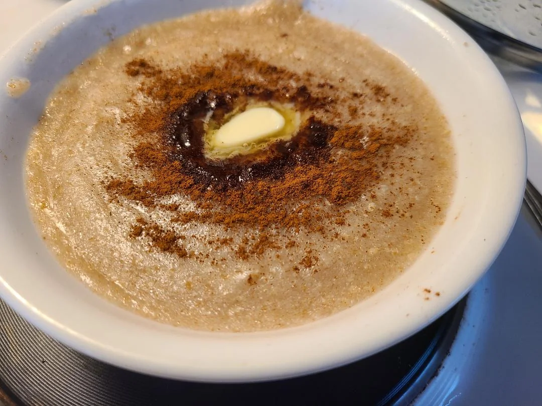 A bowl of creamy porridge topped with melted butter, a sprinkle of cinnamon, and a drizzle of syrup in the center. The bowl is white and placed on a stovetop burner.