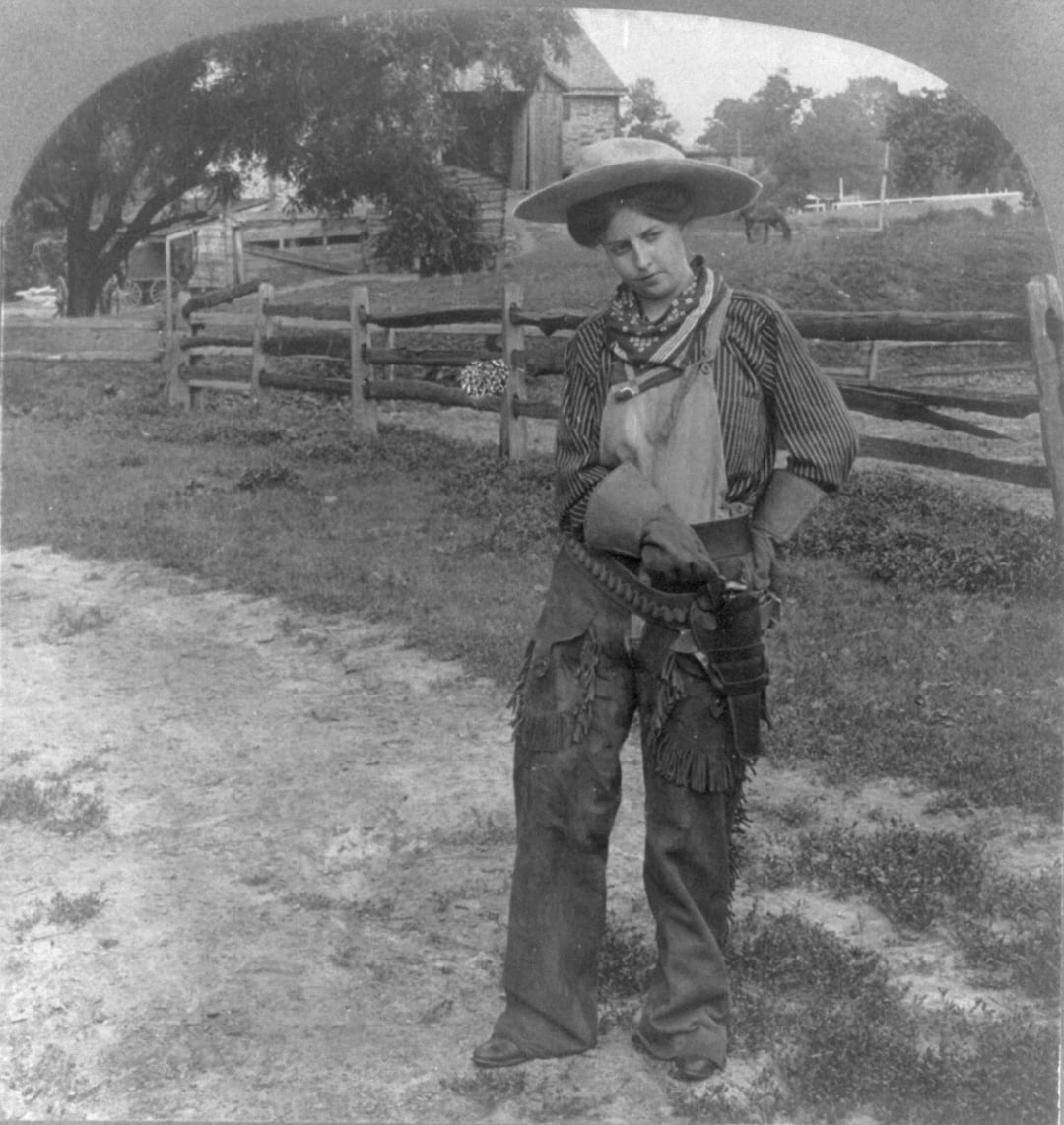A person dressed as a cowboy stands on a dirt path by a wooden fence, wearing a wide-brimmed hat, striped shirt, chaps, neckerchief, and gun holster, with a ranch and trees in the background.