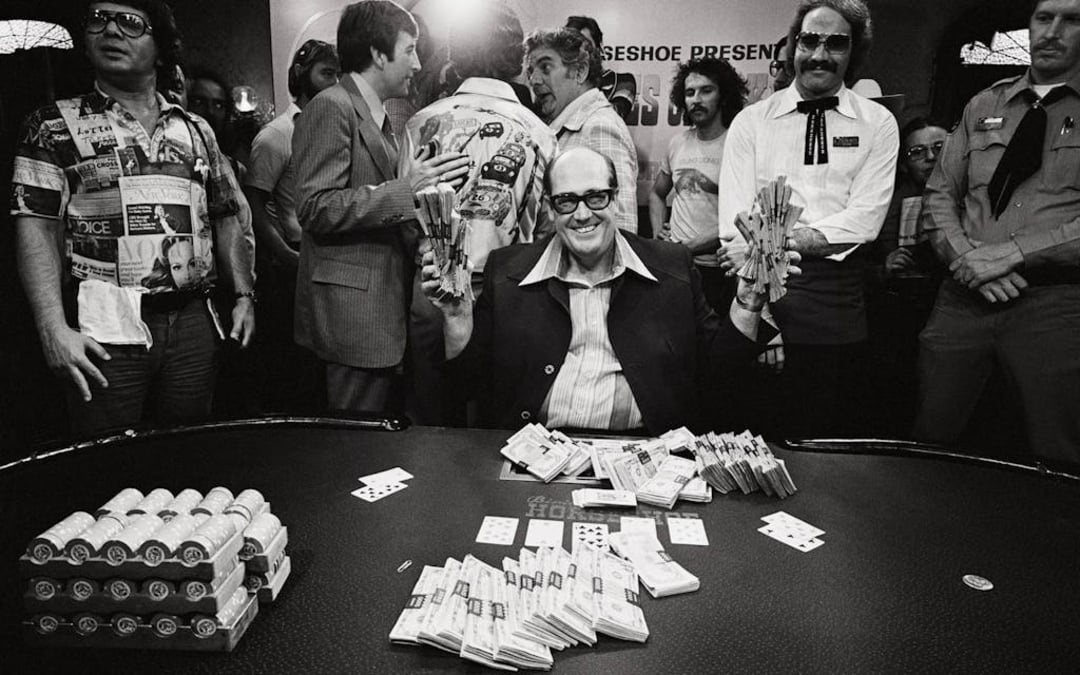 A man sits at a poker table smiling, holding bundles of cash, with large stacks of chips and money in front of him. Several people stand around, some clapping, in a crowded, lively casino atmosphere.