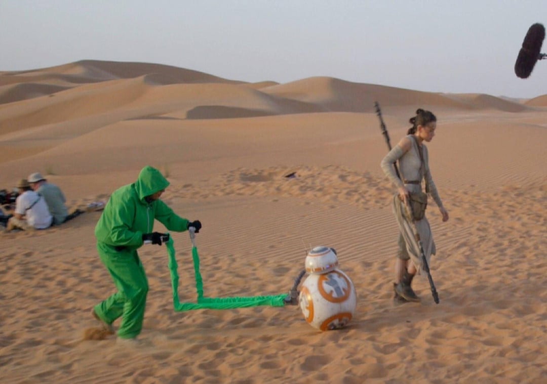 On a desert film set, a person in a green suit guides the BB-8 droid prop with a rod while an actress dressed in Star Wars-style clothing walks ahead, sand dunes and crew members visible in the background.