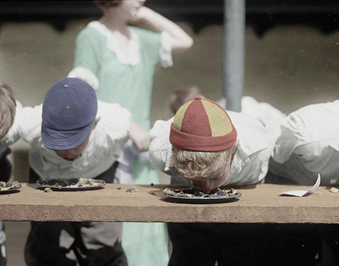 Two children, hands behind their backs, compete in a pie-eating contest outdoors, faces close to their pies on a wooden table. One wears a blue cap and the other a red and yellow cap. A woman stands in the background.