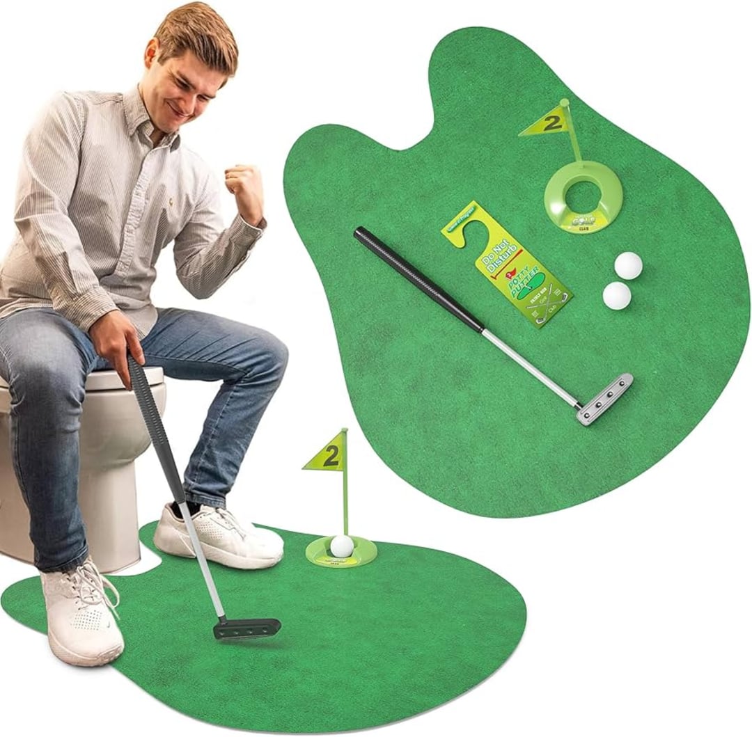 A man sitting on a toilet plays a mini golf game using a small green putting mat, club, two golf balls, and a plastic hole with a flag numbered “2.” The putting set is designed for use in the bathroom.