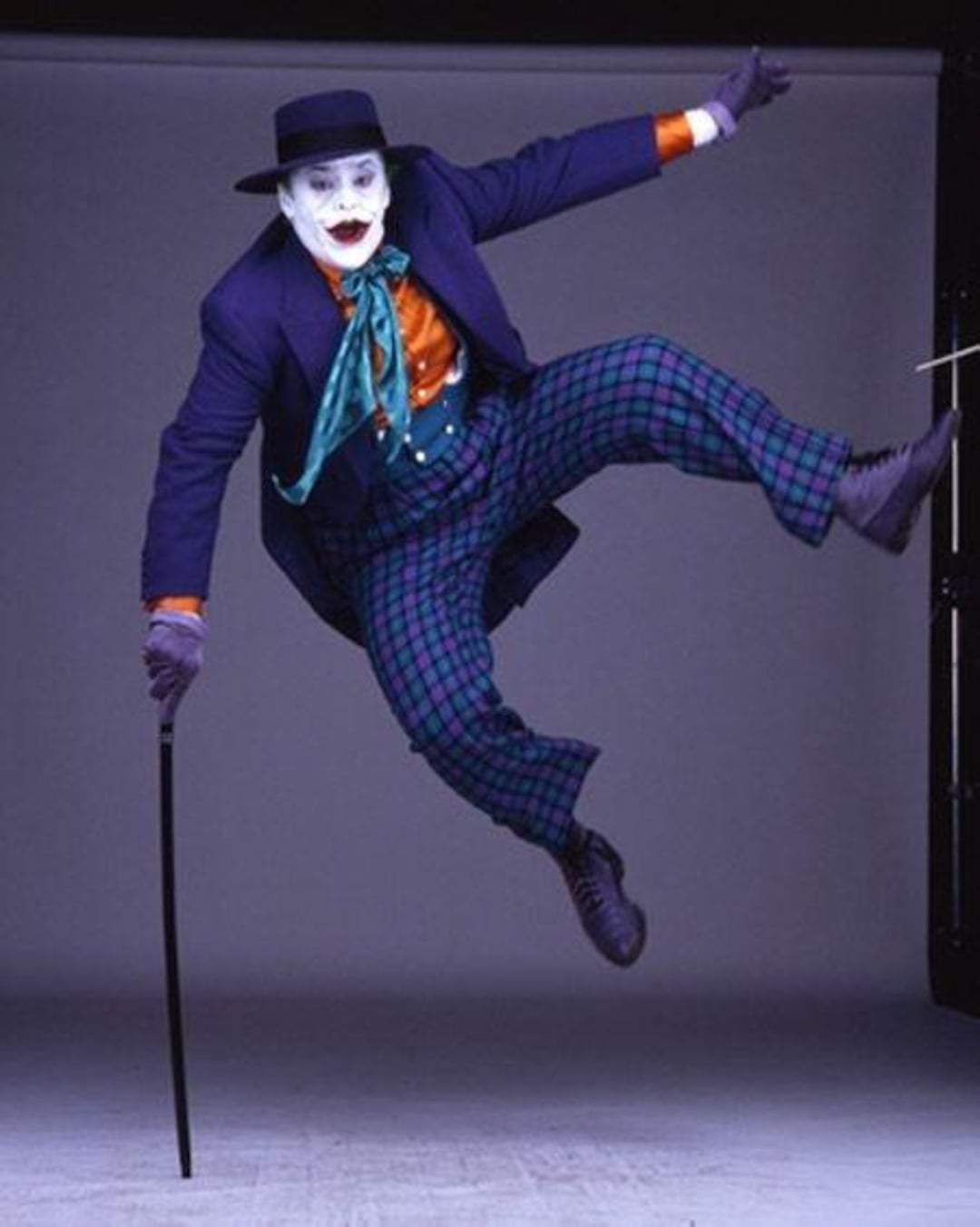 A person dressed as the Joker, wearing a purple suit, plaid pants, green bow tie, and white face makeup, jumps energetically in the air while holding a cane.