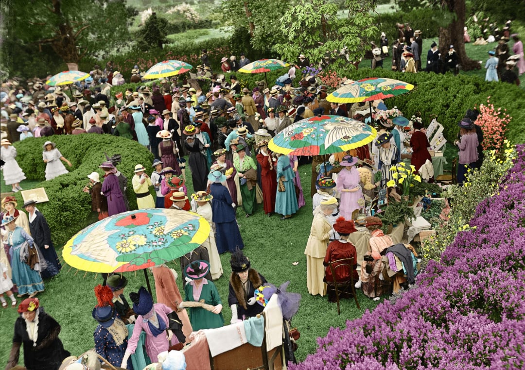 A large outdoor garden party with many women in colorful early 20th-century dresses and hats, standing under bright, decorated parasols, surrounded by lush greenery and purple flowers.