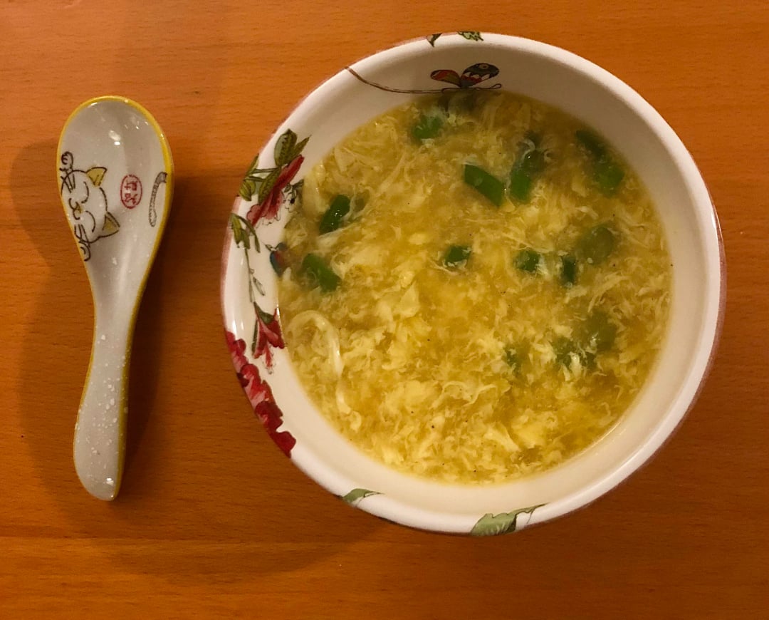 A bowl of egg drop soup with green onions, placed on a wooden surface next to a decorative ceramic spoon with a cat design.