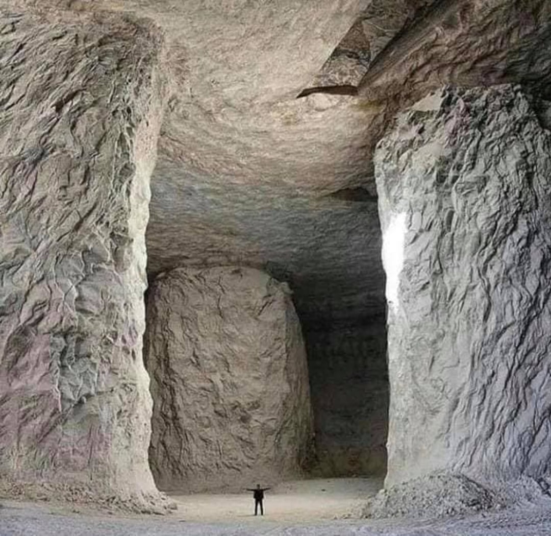 A person stands with arms raised inside a massive cave or cavern with towering rock pillars, rough textured walls, and a high ceiling, emphasizing the immense scale of the underground space.