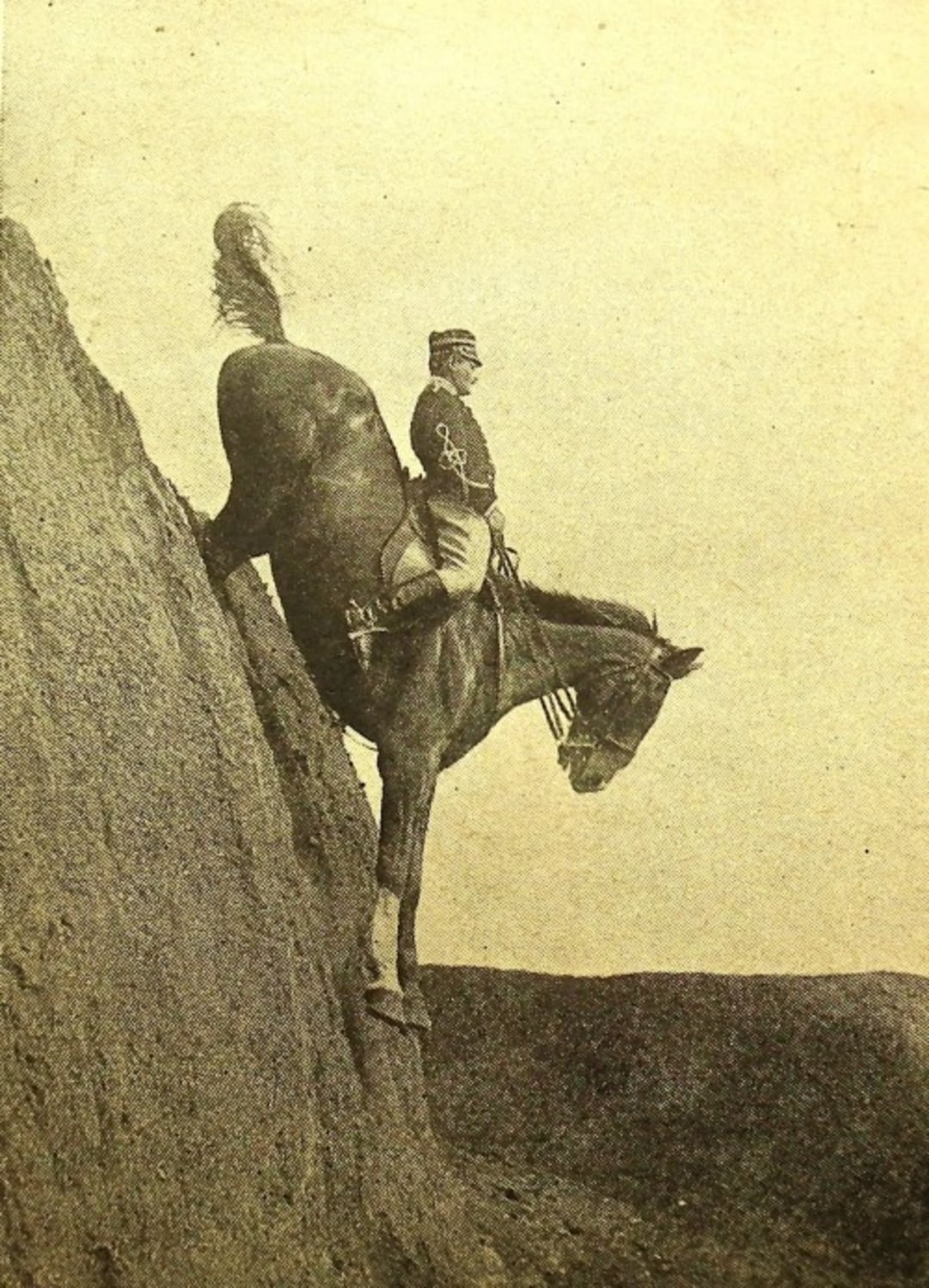 A soldier in uniform rides a horse down a steep, nearly vertical rocky slope. The horse is angled sharply downward, with its front legs extended and tail raised, while the rider sits upright in the saddle.