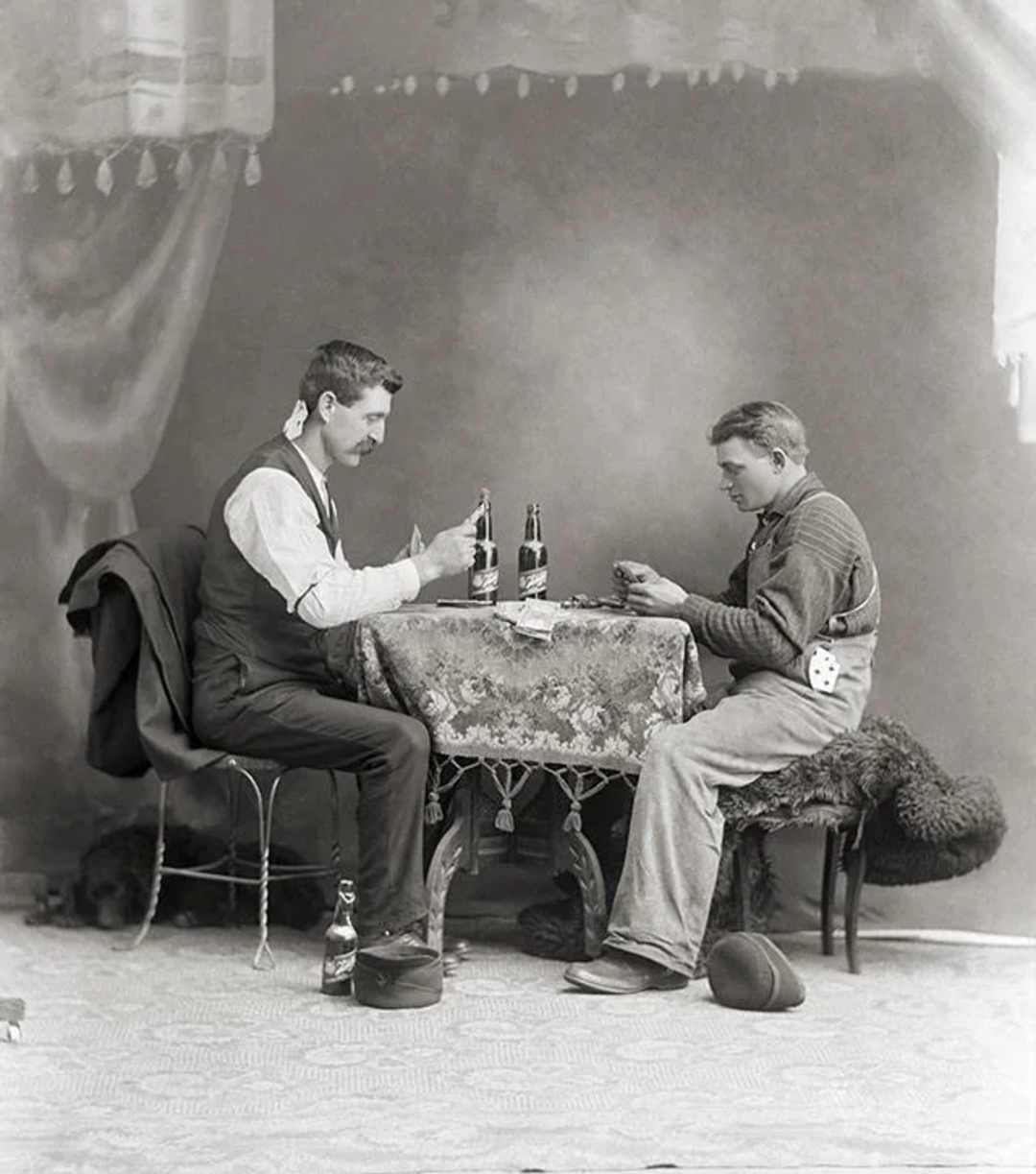 Two men sit across from each other at a small, ornate table with a patterned cloth, playing cards. Bottles and glasses are on the table; hats rest on the floor. The scene appears vintage, with a curtain backdrop.