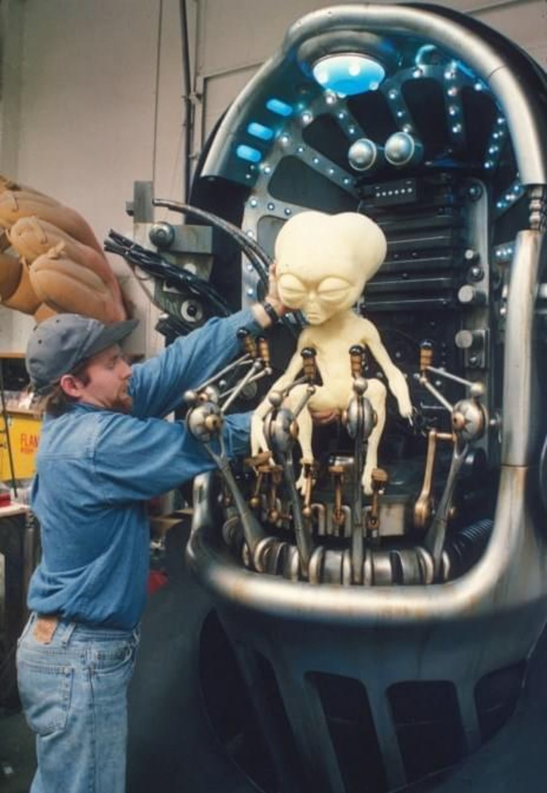 A person in a denim shirt and cap works on a large animatronic alien figure with a big head, seated in a futuristic, metallic control chair surrounded by mechanical arms and blue lights.