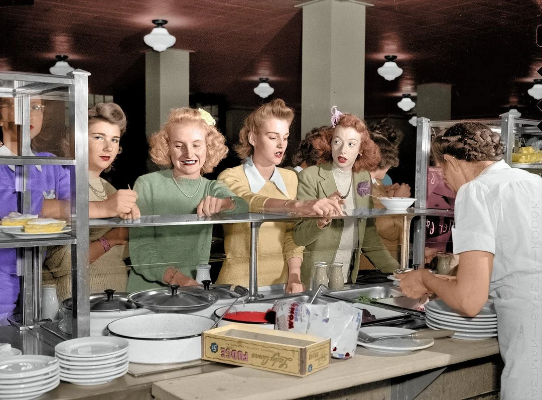 A group of young women in vintage clothing stand in line at a cafeteria counter, holding trays and waiting to be served food by a worker in white attire. The setting appears to be from the mid-20th century.