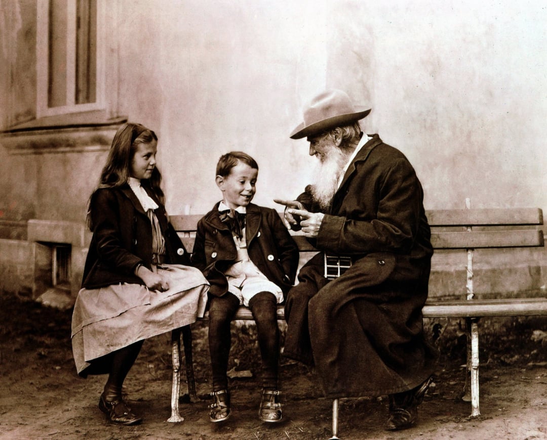 An elderly man with a long beard sits on a bench, showing an object to a smiling boy and a girl. All three wear old-fashioned clothes. The scene appears to be outdoors, near a building wall.