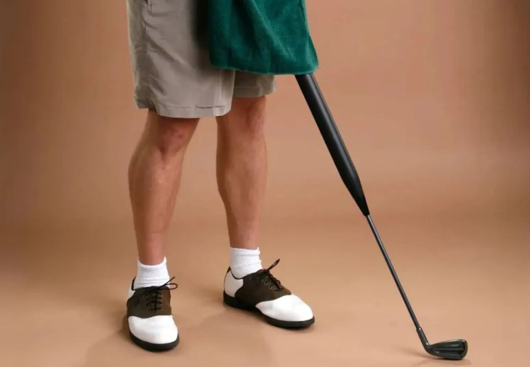A person wearing shorts, white socks, and golf shoes stands on a tan floor. Instead of a right leg, there is a prosthetic shaped like a golf club.