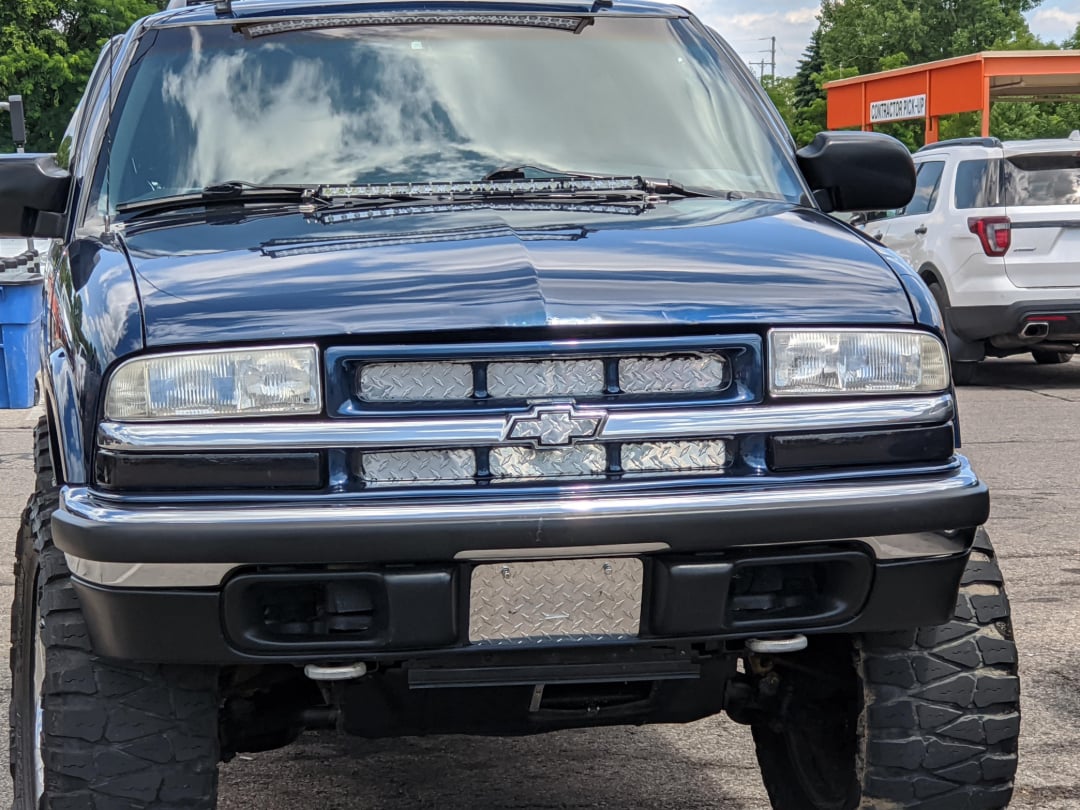 A lifted, dark blue Chevrolet SUV is parked on the street. The front grille and bumper have diamond plate detailing, and the vehicle has large off-road tires. Other cars and trees are visible in the background.