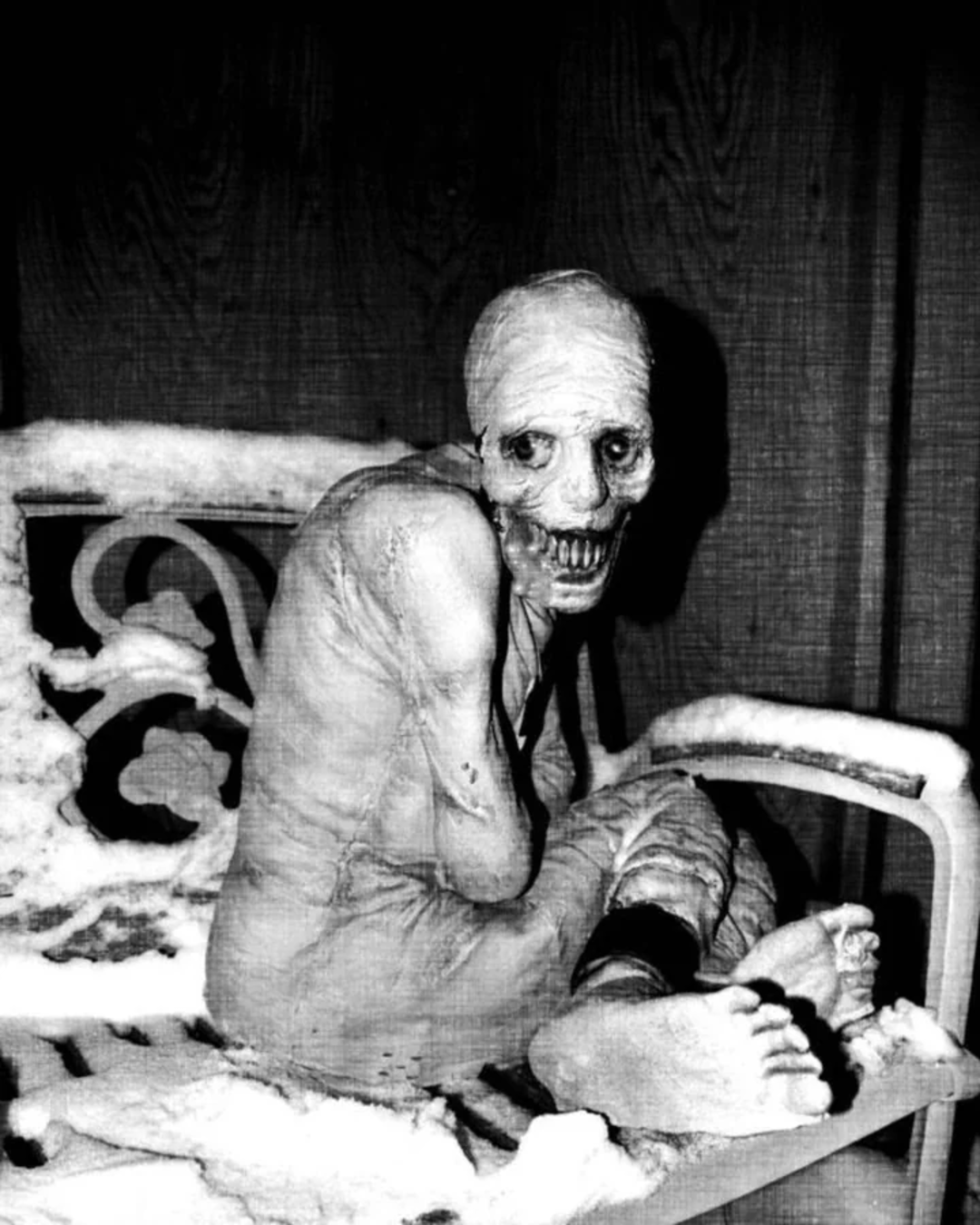 A skeletal, mummy-like figure with hollow eyes and a wide, toothy grin sits hunched on a snow-covered bench in a dark, eerie setting.