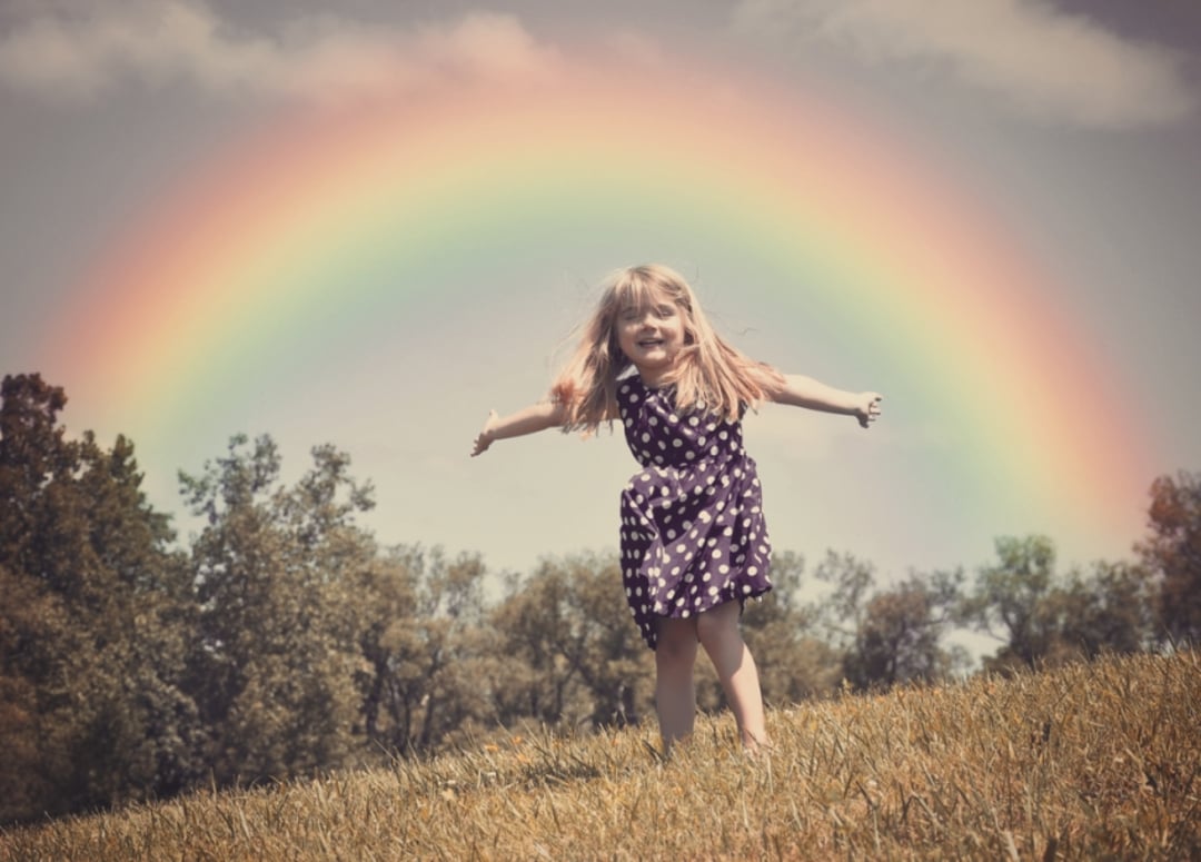 A young girl in a polka dot dress stands on a grassy hill with arms outstretched, smiling joyfully. A bright, colorful rainbow arches across the sky behind her, and trees are visible in the background.