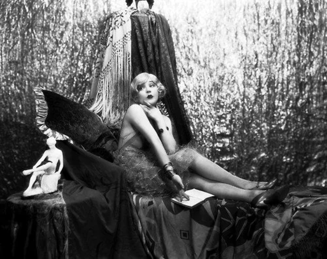 A woman in vintage 1920s-style clothing lounges on an ornate, draped chaise, surrounded by decorative fabrics and a small statue, against a shimmering, textured backdrop. She gazes thoughtfully to the side.